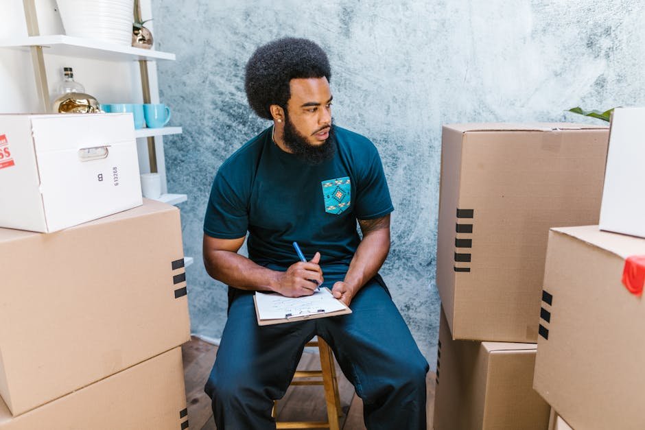 A focused male mover sits with a clipboard amid cardboard boxes, planning relocation tasks.