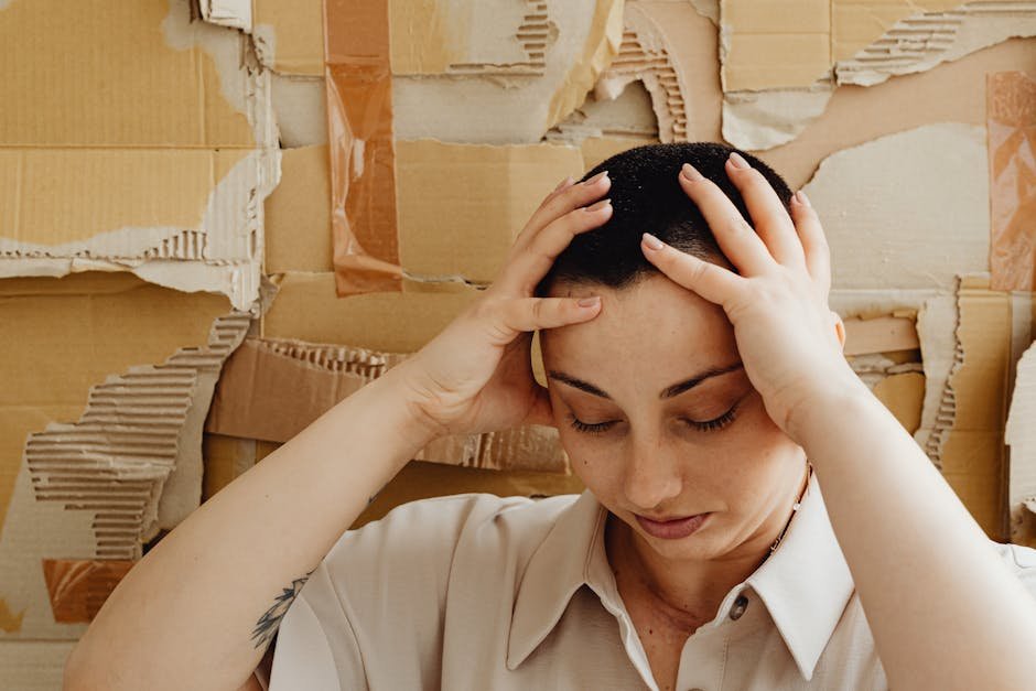 A contemplative woman with a buzz cut rests her hands on her head against a textured cardboard background.